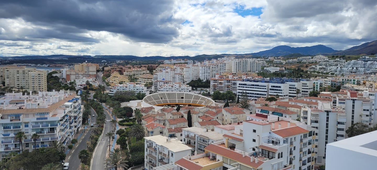 Elevated panoramic view of Estepona near LAS MESAS BLUE HORIZON, featuring the Plaza de Toros bullring, white-washed residential buildings, a palm-lined boulevard and the Sierra Bermeja mountain backdrop.