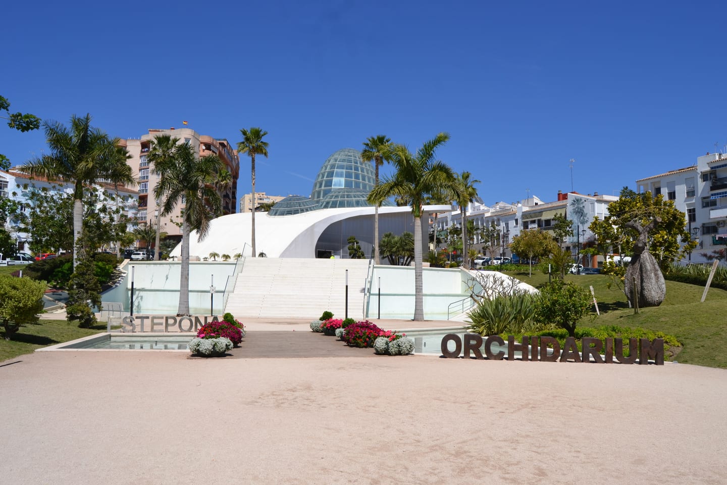 INSUR SCALA — Area photograph: Estepona Orchidarium with its glass dome, white curved architecture, palm trees and gardens.
