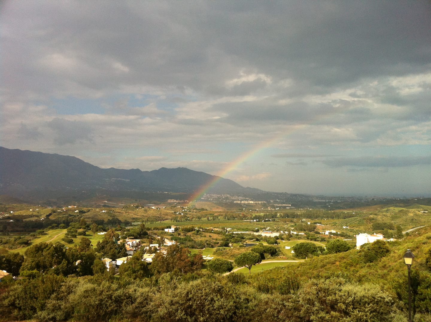 Panoramic valley view of the La Cala Golf / Mijas countryside with golf course fairways, scattered white villas, Mediterranean scrubland, mountain backdrop, and a rainbow under a dramatic sky, shown as local context near LA RESERVA DE LA CALA GOLF
