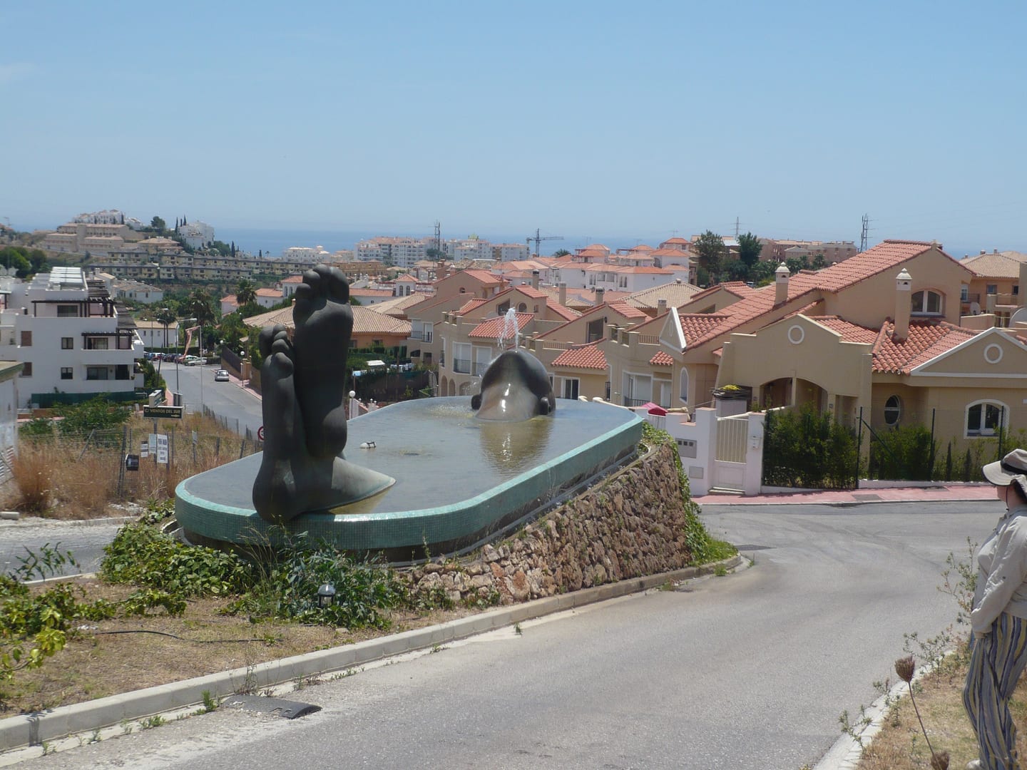 Area photograph near BOUTIQUE PROJECT showing a public fountain sculpture with abstract figures in a roundabout, with the La Cala de Mijas hillside residential townscape and Mediterranean Sea behind.