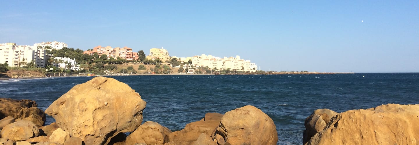 Rocky Mediterranean shoreline with Estepona's waterfront residential skyline across a calm bay near NATURA ESTEPONA