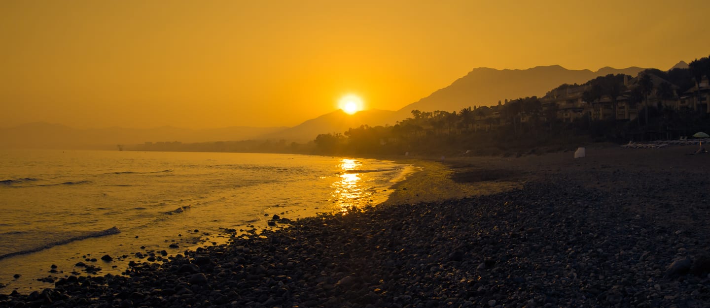 Sunset over the Marbella Mediterranean coastline with Sierra Blanca mountain silhouette and coastal residential hillside, shown as local context near THE LIST RIO REAL
