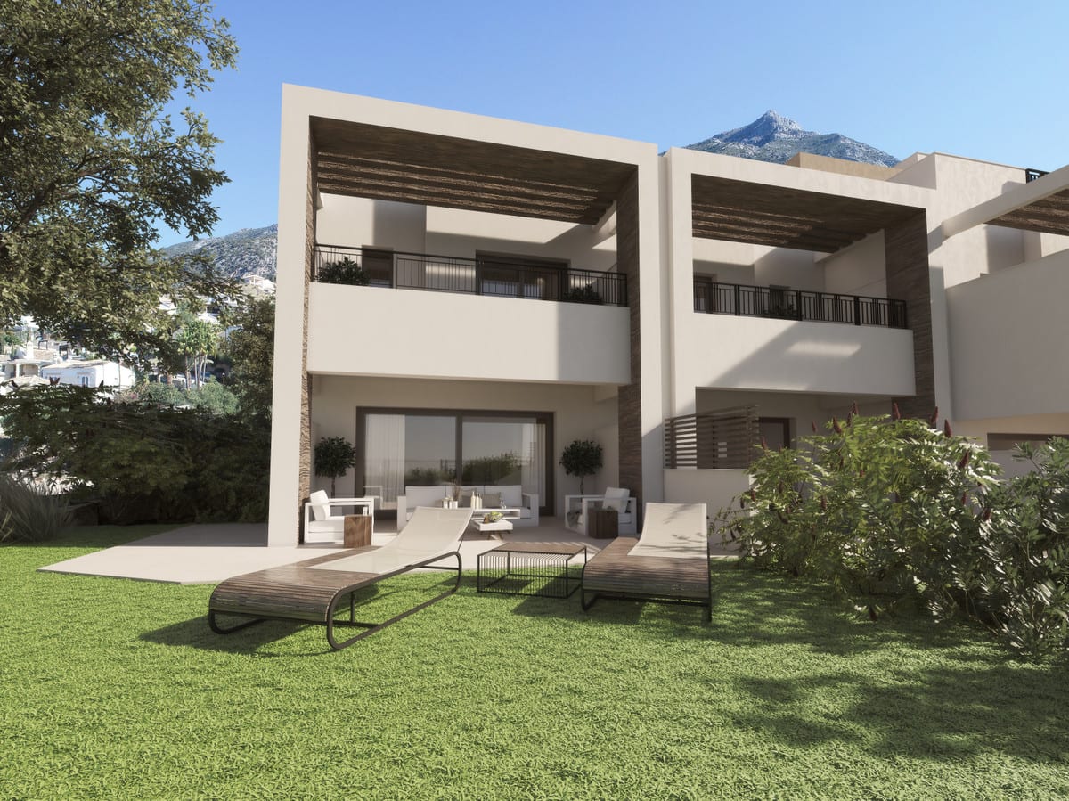 Project visualisation of ALMAZARA VIEWS (TOWNHOUSES) featuring a green lawn, two sun loungers, a covered outdoor area with seating and sand-beige façades with dark balustrades.
