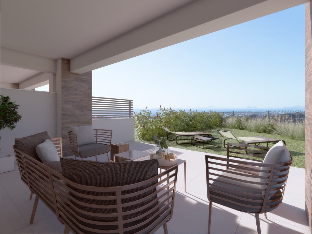 Project visualisation of ALMAZARA VIEWS (TOWNHOUSES) featuring a covered terrace, chairs with woven seats and metal frames, an adjoining lawn with sun loungers and a distant view of water on the horizon.