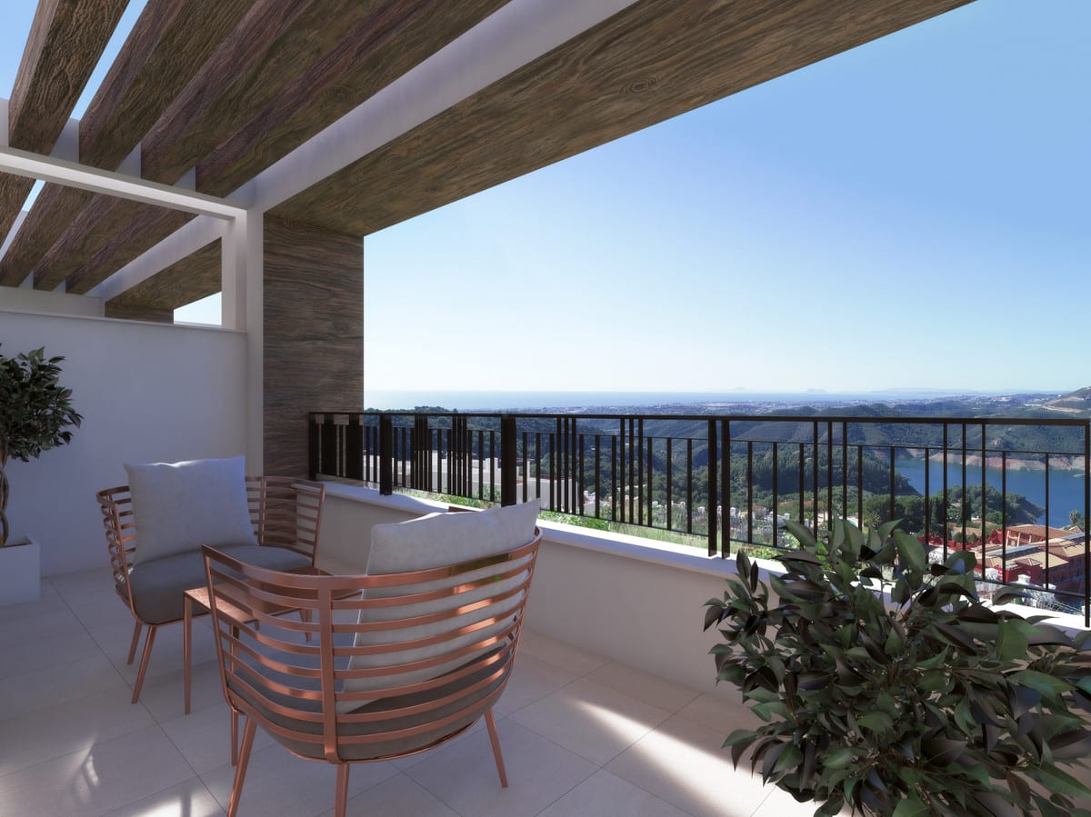 Project visualisation of ALMAZARA VIEWS (TOWNHOUSES) featuring a terrace beneath timber beams, a dark metal balustrade, armchairs with copper-coloured frames and a broad landscape of hills and water in the background.