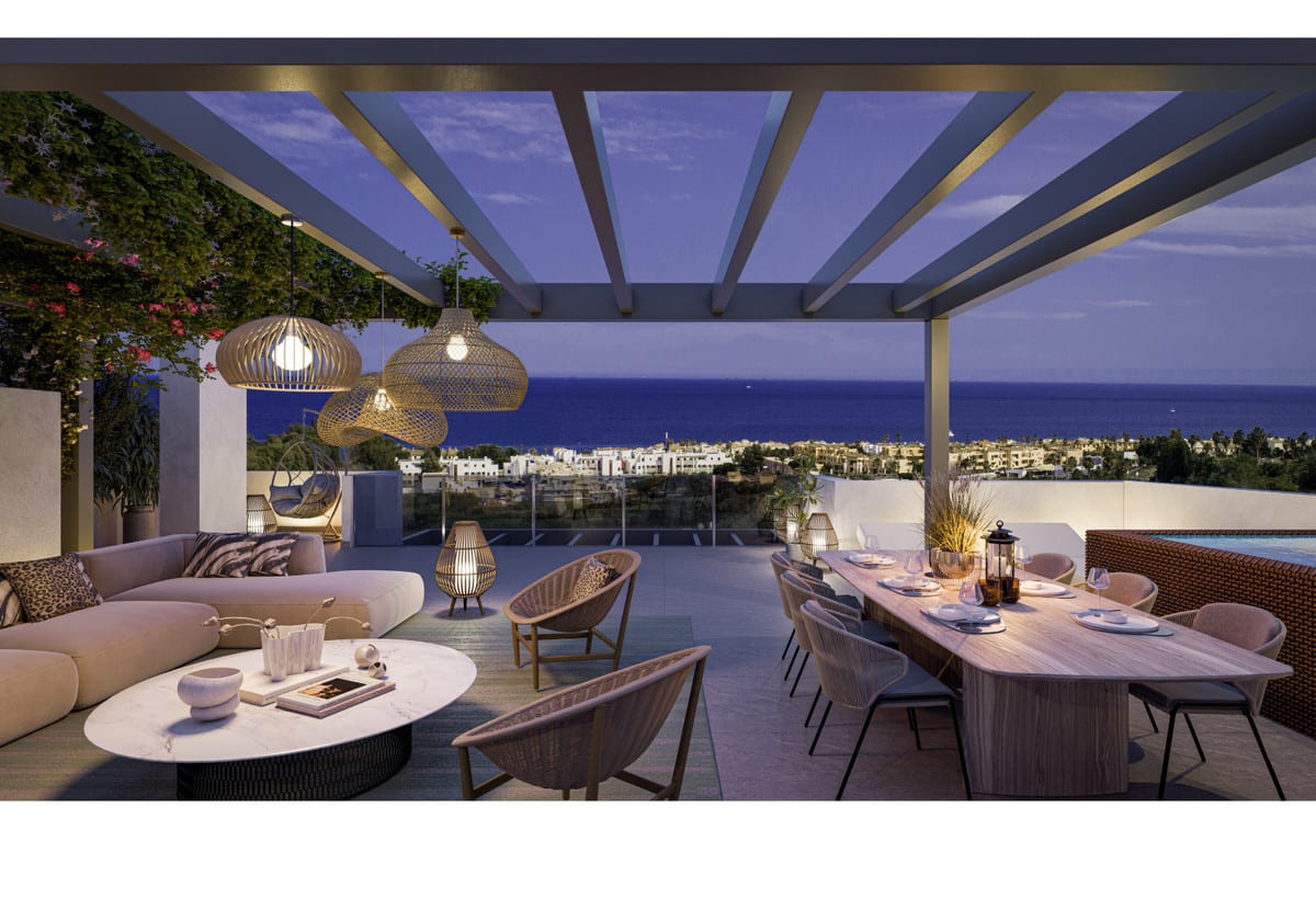 Rendered evening visualisation of ALURA LIVING with a covered terrace beneath a pergola, woven pendant lights, lounge area with a curved sofa and armchairs, wooden dining table with chairs, lanterns and views of the sea and surrounding buildings at dusk.