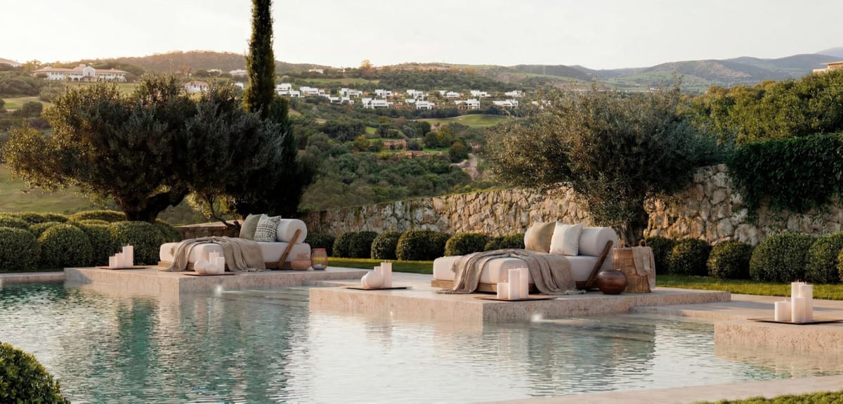 Project visualisation of AMENÁBAR CORTESÍN with lounge beds on stone platforms in the swimming pool, candles, olive trees, rounded shrubs and a natural stone wall in the background.