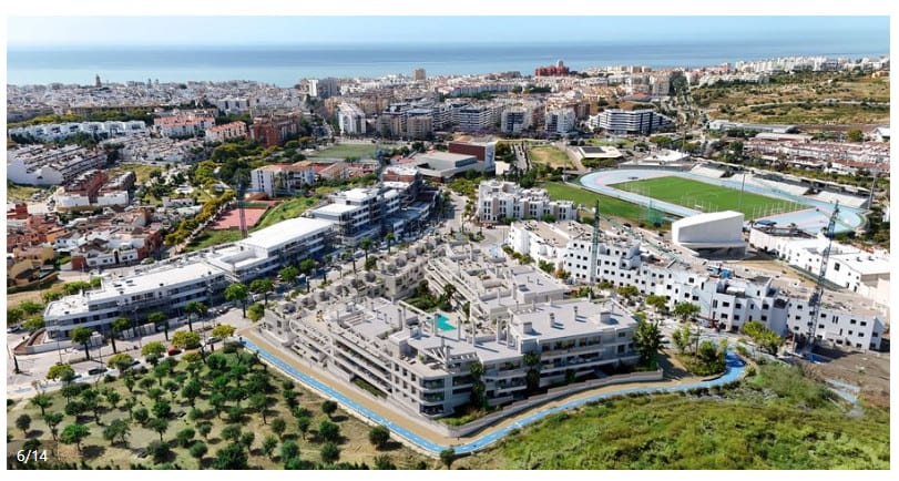 Aerial photograph with a project visualisation of ANNA DE ESTEPONA, showing the rendered building volume under construction, surrounding streets, trees, an athletics track and urban buildings in the background.