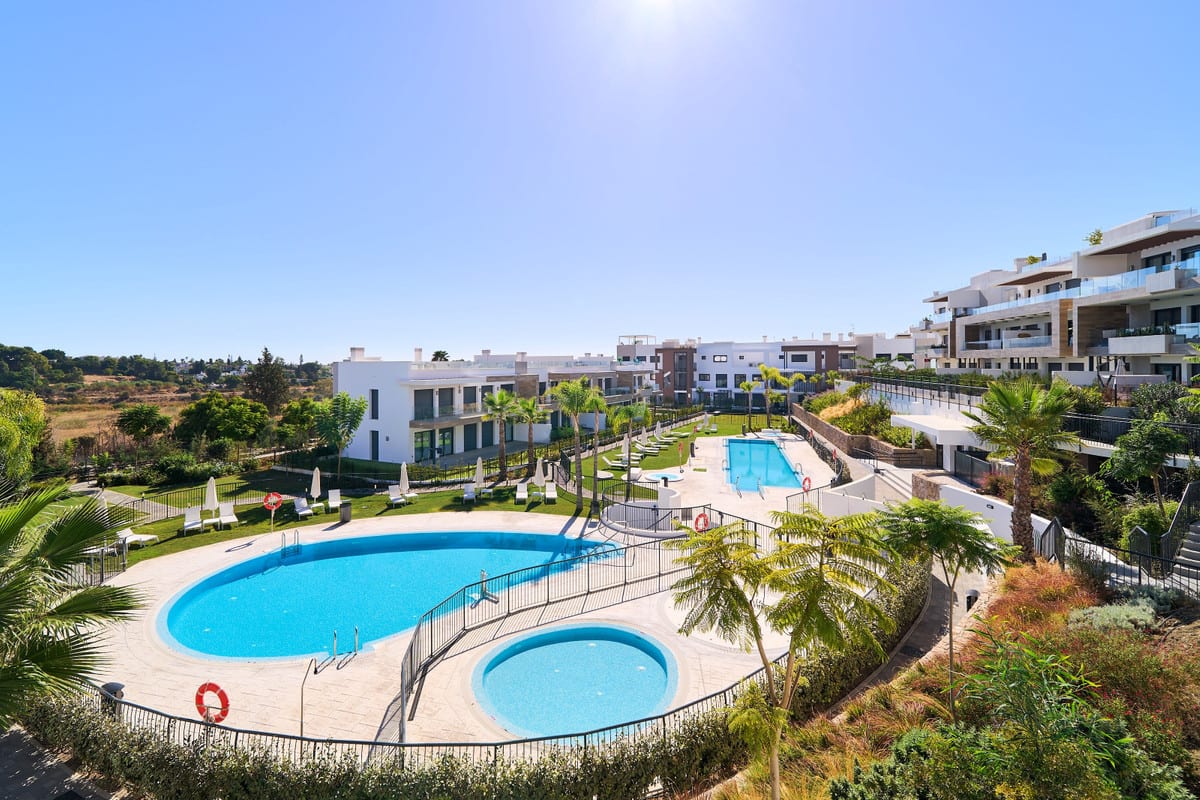 ATALAYA EMOTION with an organically shaped swimming pool and a smaller round pool on a light stone terrace, surrounded by black fencing, planting, sun loungers, closed parasols and white buildings.