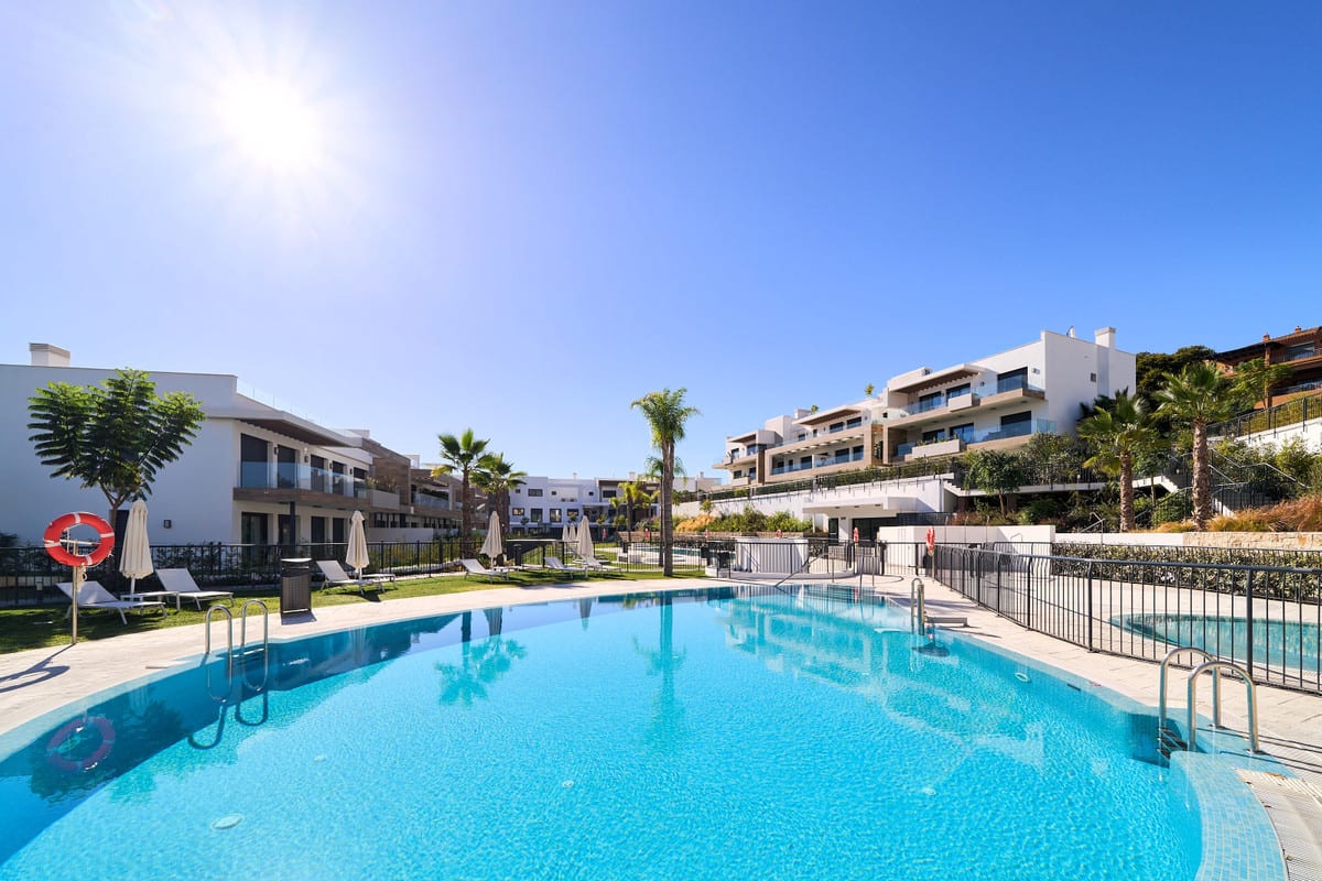 ATALAYA EMOTION with a large organically shaped swimming pool in the foreground, metal handrails, sun loungers, closed parasols, palm trees and white buildings with balconies on a slope.