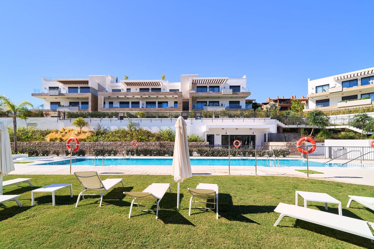 ATALAYA EMOTION with white sun loungers on a lawn in the foreground, a closed parasol, a long rectangular swimming pool with lifebuoys and white buildings with stone cladding and balconies in the background.