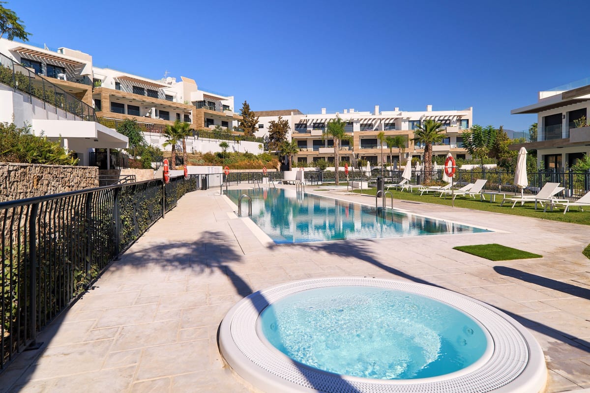 ATALAYA EMOTION with a round hot tub on a light stone terrace in the foreground, a long rectangular swimming pool, black metal fencing, palm trees, sun loungers, parasols and white buildings with stone cladding and balconies.