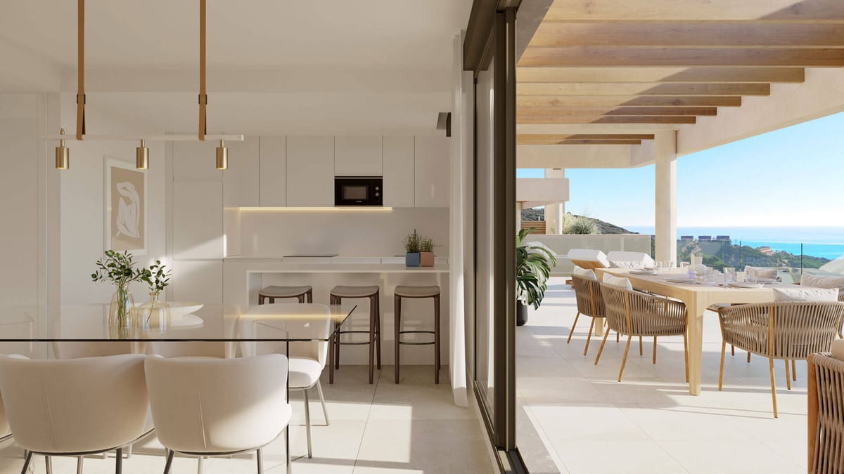 Rendered interior visualisation of ATRIA - PHASE 1 with a white kitchen, glass dining table with chairs, kitchen island with bar stools and a covered terrace with wooden beams, an outdoor dining area and sea views.