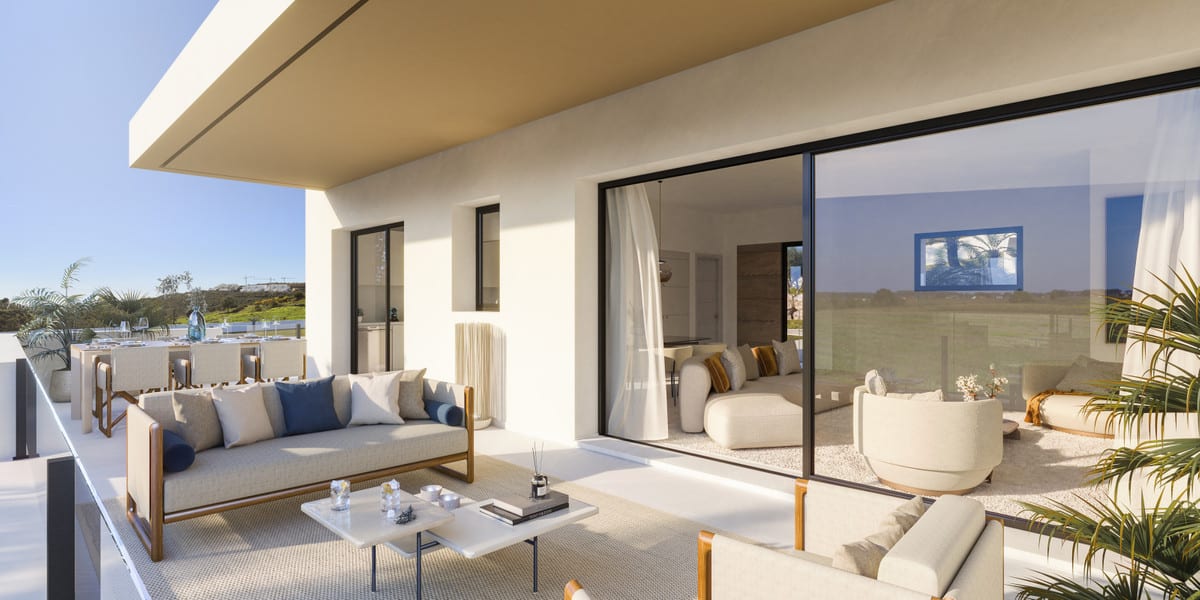 Project visualisation of ATTIRE ESTEPONA featuring a furnished terrace, two lounge sofas with beige and blue cushions, a marble coffee table and large black-framed sliding glass doors leading to the interior.