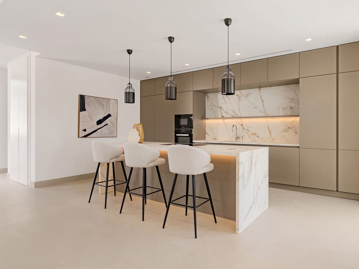 Kitchen island with a marble-effect finish, light bar stools and three pendant lights against taupe cabinets.