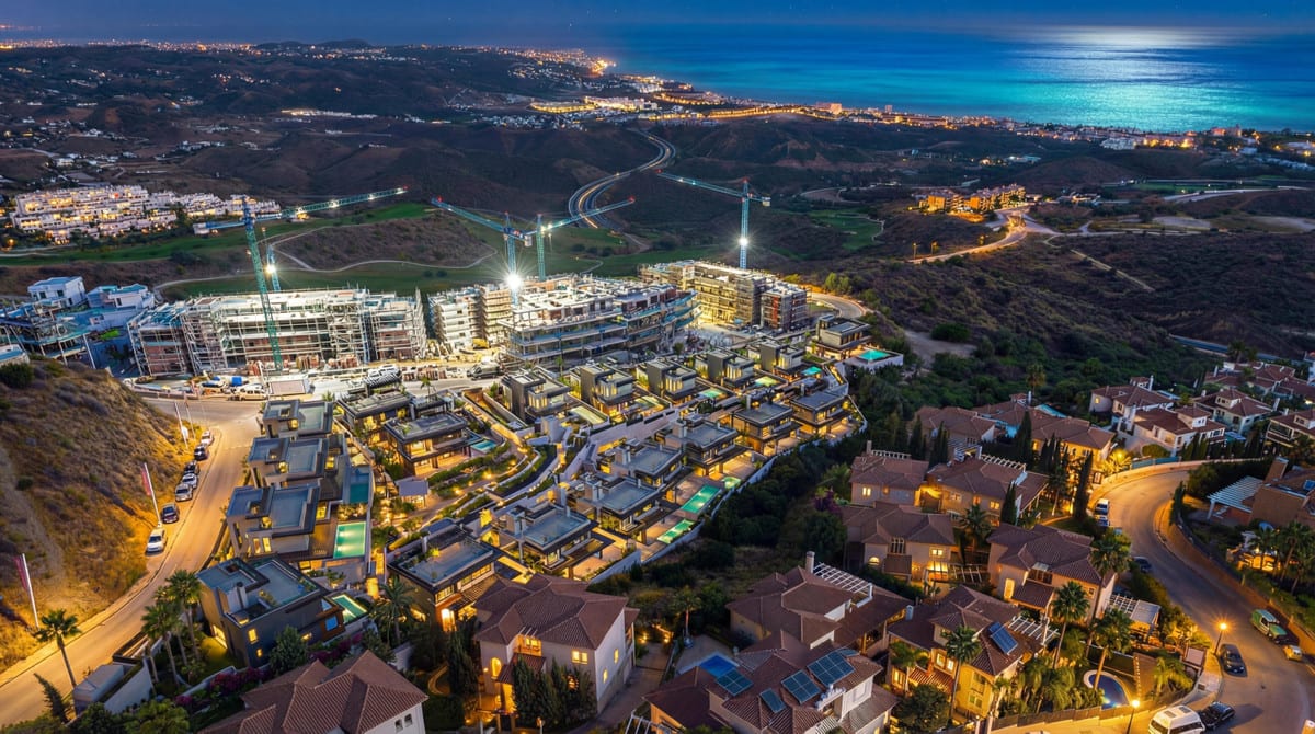 Dusk aerial photograph of BALI VILLAS under construction, with construction cranes, illuminated streets, surrounding hills and the sea on the horizon.