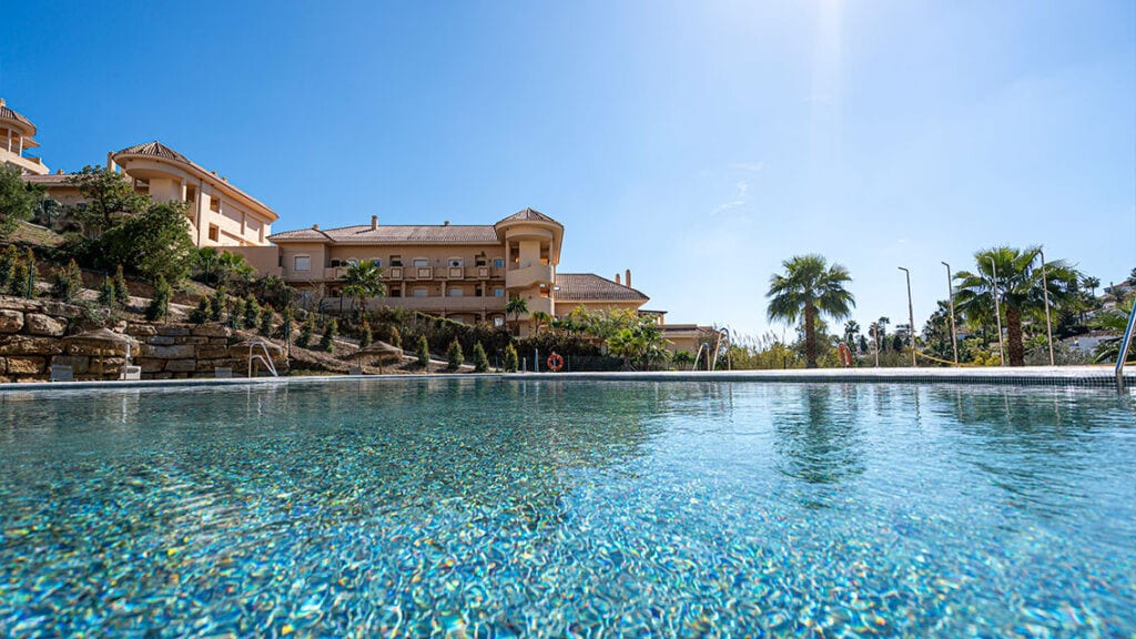 BE ALOHA with large swimming pool, palm trees and a sand-coloured building with a terracotta roof beneath a blue sky