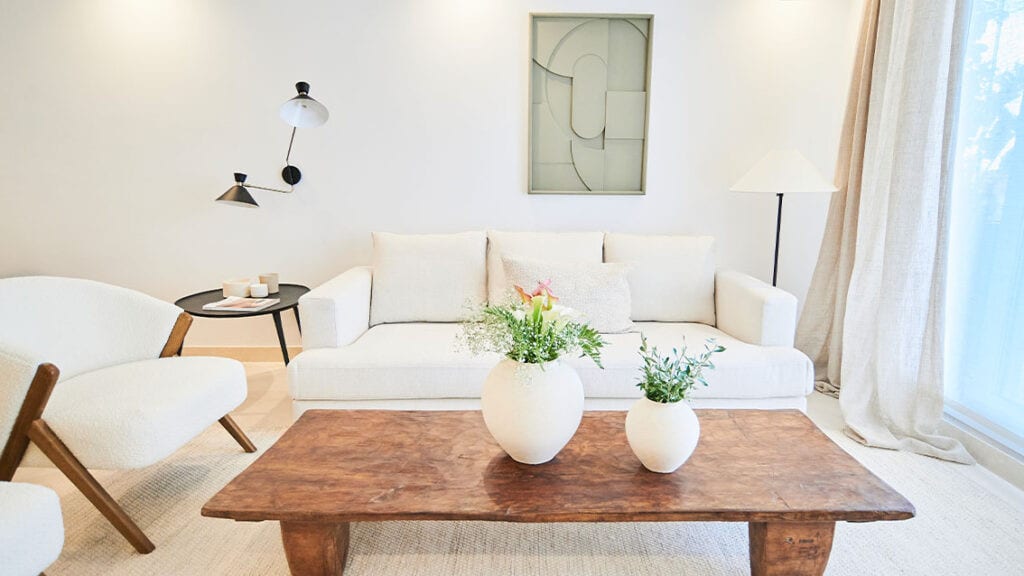 BE ALOHA seating area with white sofa, wooden coffee table, an armchair, vases of flowers and a linen curtain