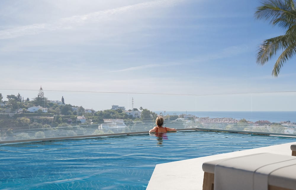BELVEDERE COLLECTION: a person in a swimming pool behind a glass balustrade, with buildings, palm trees and the sea in the background.