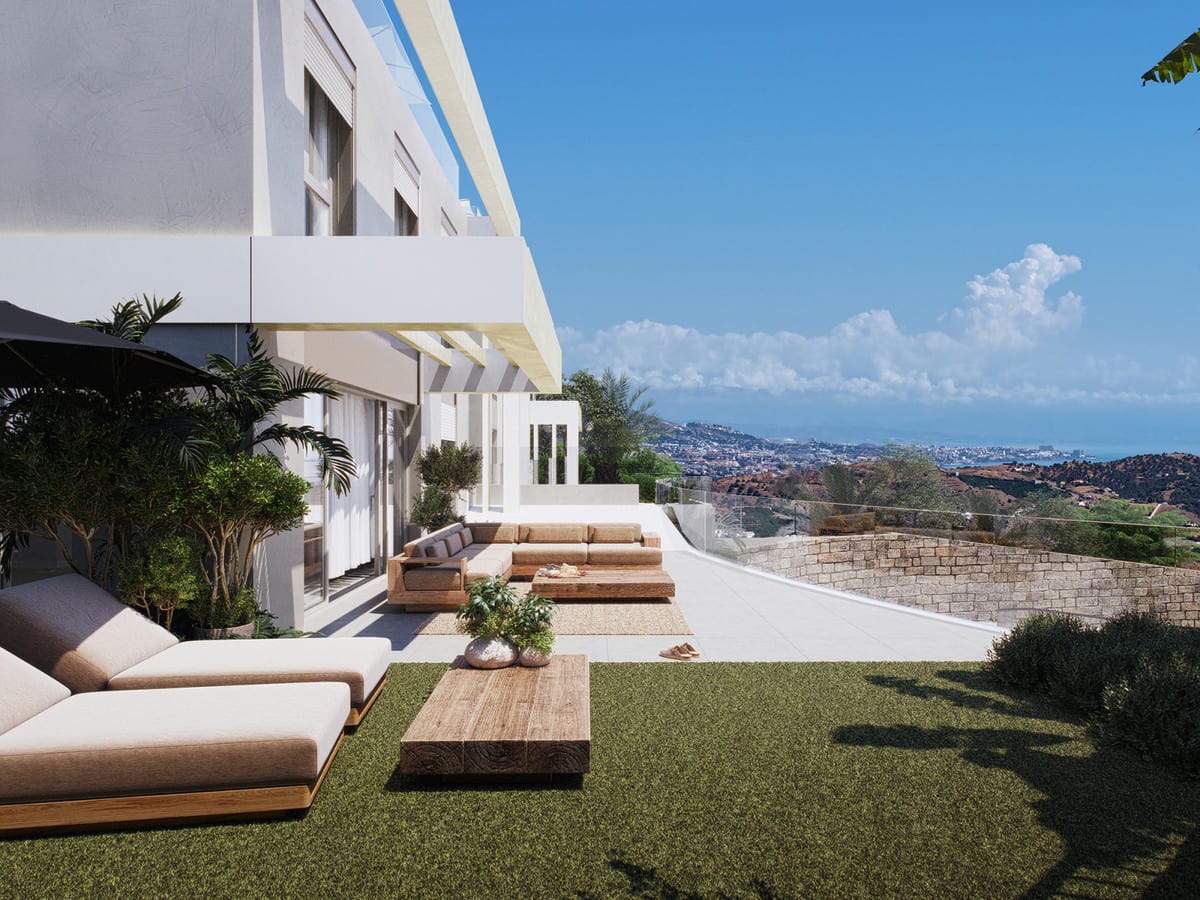 Project visualisation of CALANOVA HOMES featuring a spacious outdoor terrace beside a white façade, wooden lounge furniture, a green area, a wooden coffee table, glass balustrade and wide views over buildings, hills and the sea in the background.
