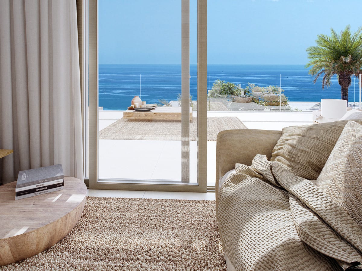 Project visualisation of CALANOVA HOMES featuring a beige armchair with woven cushions, a round wooden side table, stacked architecture books, a deep-pile rug and a glass sliding door opening onto a terrace with sea views.
