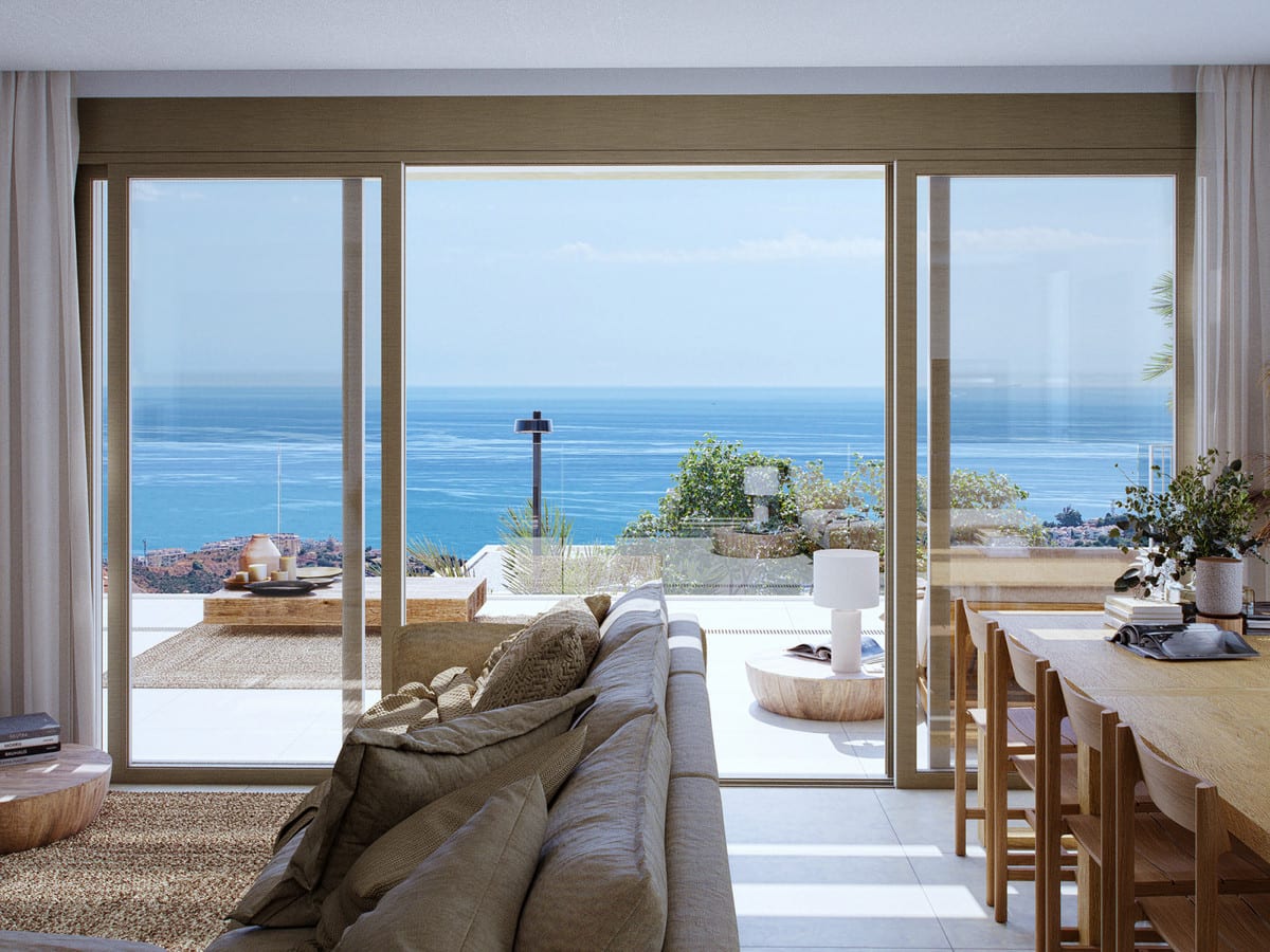 Project visualisation of CALANOVA HOMES featuring a grey-beige sofa, wooden dining table with chairs, wide three-panel glass sliding doors and a terrace with views of the sea and coastline.