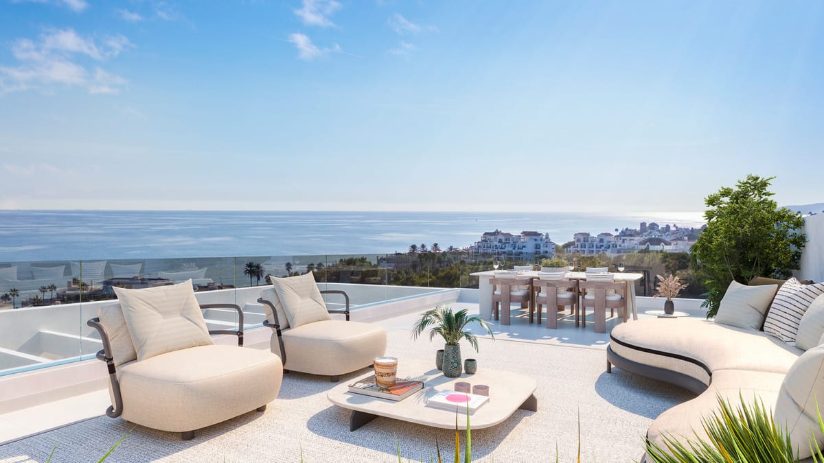Project visualisation of CELESTIA HOMES featuring a terrace with a glass balustrade, two armchairs, a curved sofa, low coffee table and round dining table with chairs, overlooking the sea and coastal buildings.