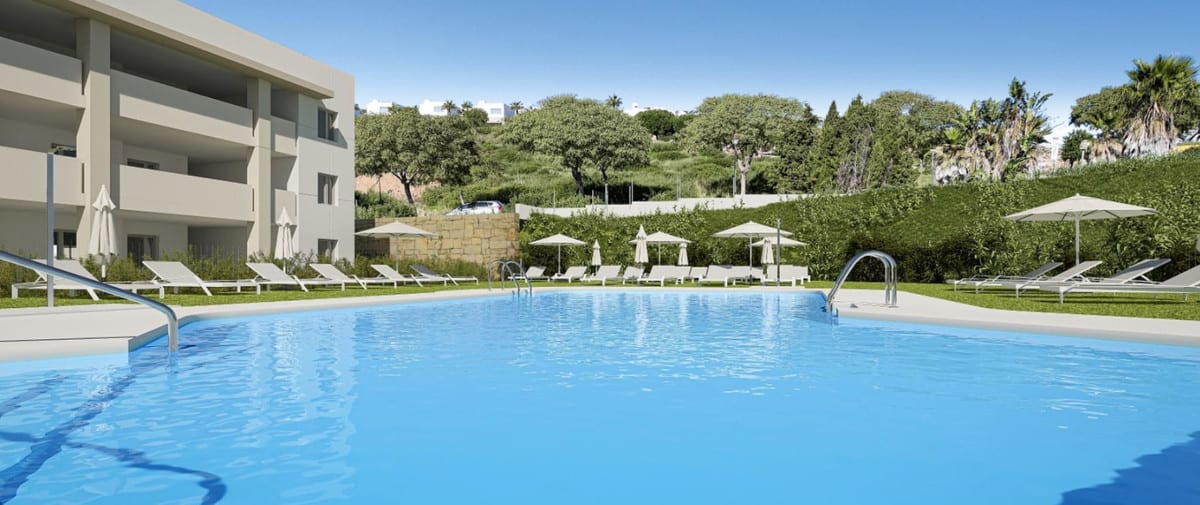 Project visualisation of GRAND BAY APARTMENTS viewed from the swimming pool, with sun loungers and parasols on the grass, a building with balconies on the left and a green hill in the background.