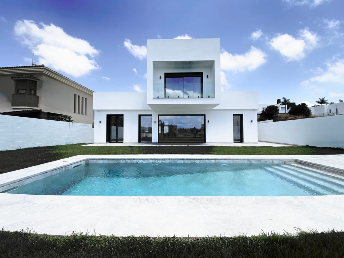 Garden view of the white villa with a rectangular pool, lawn and an upper glass balustrade.