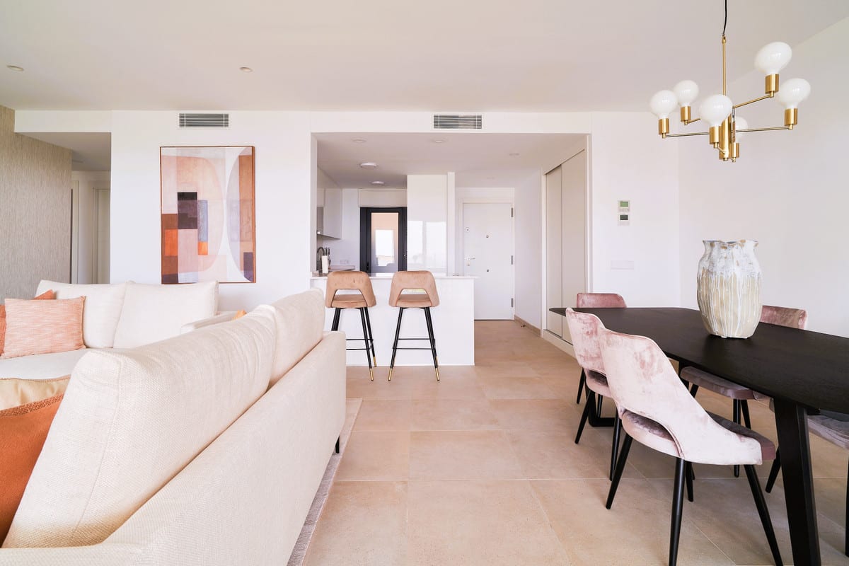 HIGUERÓN BAY RESIDENCES with a cream corner sofa with decorative cushions, a black dining table with pink velvet chairs, a white kitchen island with two taupe bar stools and a gold pendant light.