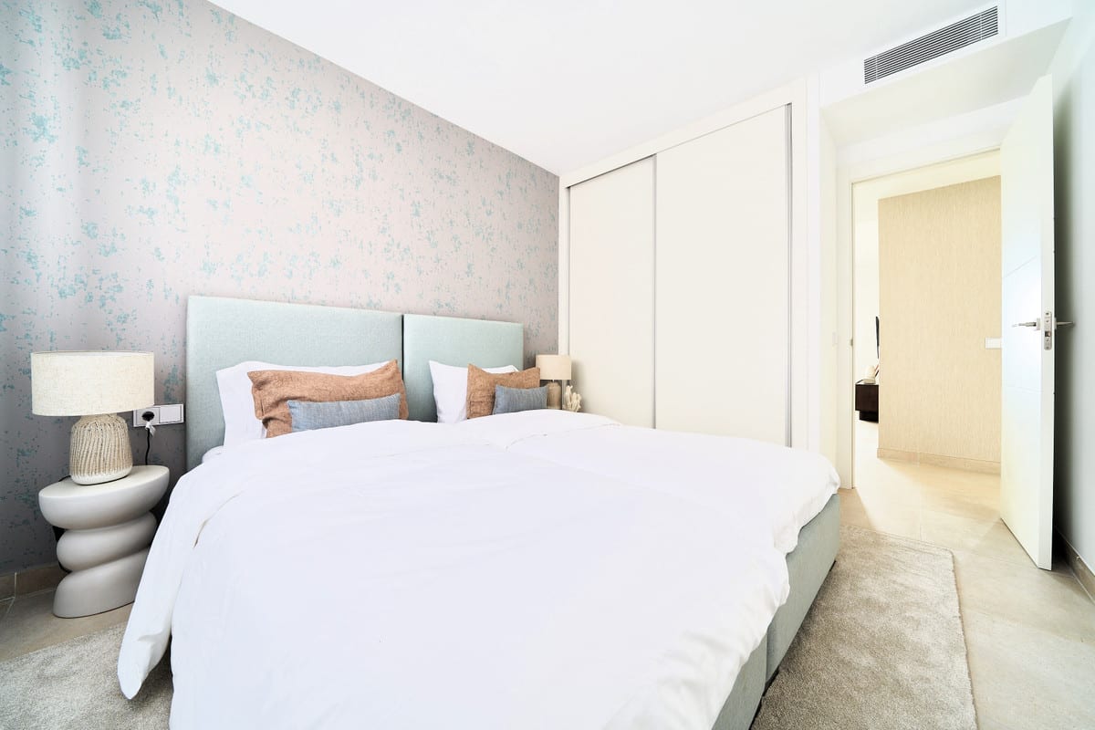 HIGUERÓN BAY RESIDENCES with a double bed, a light blue upholstered headboard in two panels, a sculptural bedside table with lamp, a cream fitted sliding wardrobe and wallpaper with a light blue pattern.
