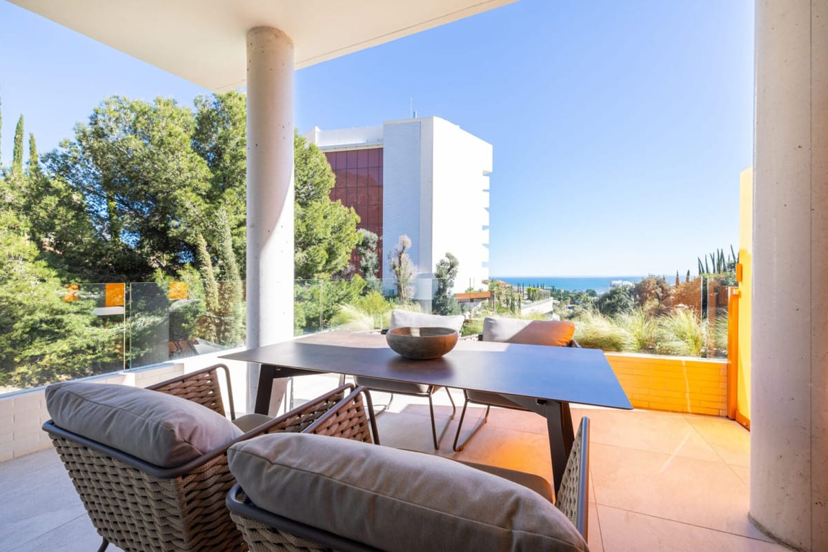 View of a covered dining terrace with woven chairs, planting and the sea in the distance.