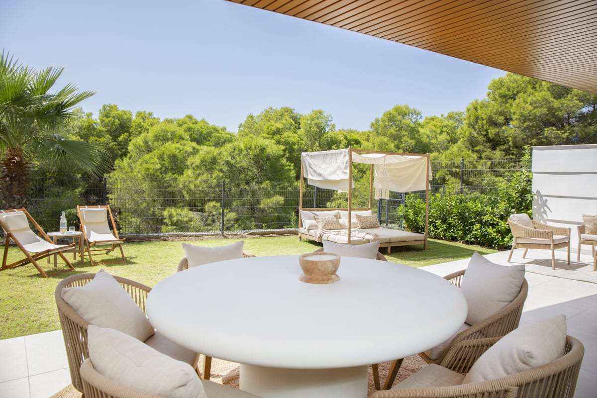 HIGUERON WEST with a round white outdoor table on a covered terrace, lawn and wooden daybed with a fabric canopy.