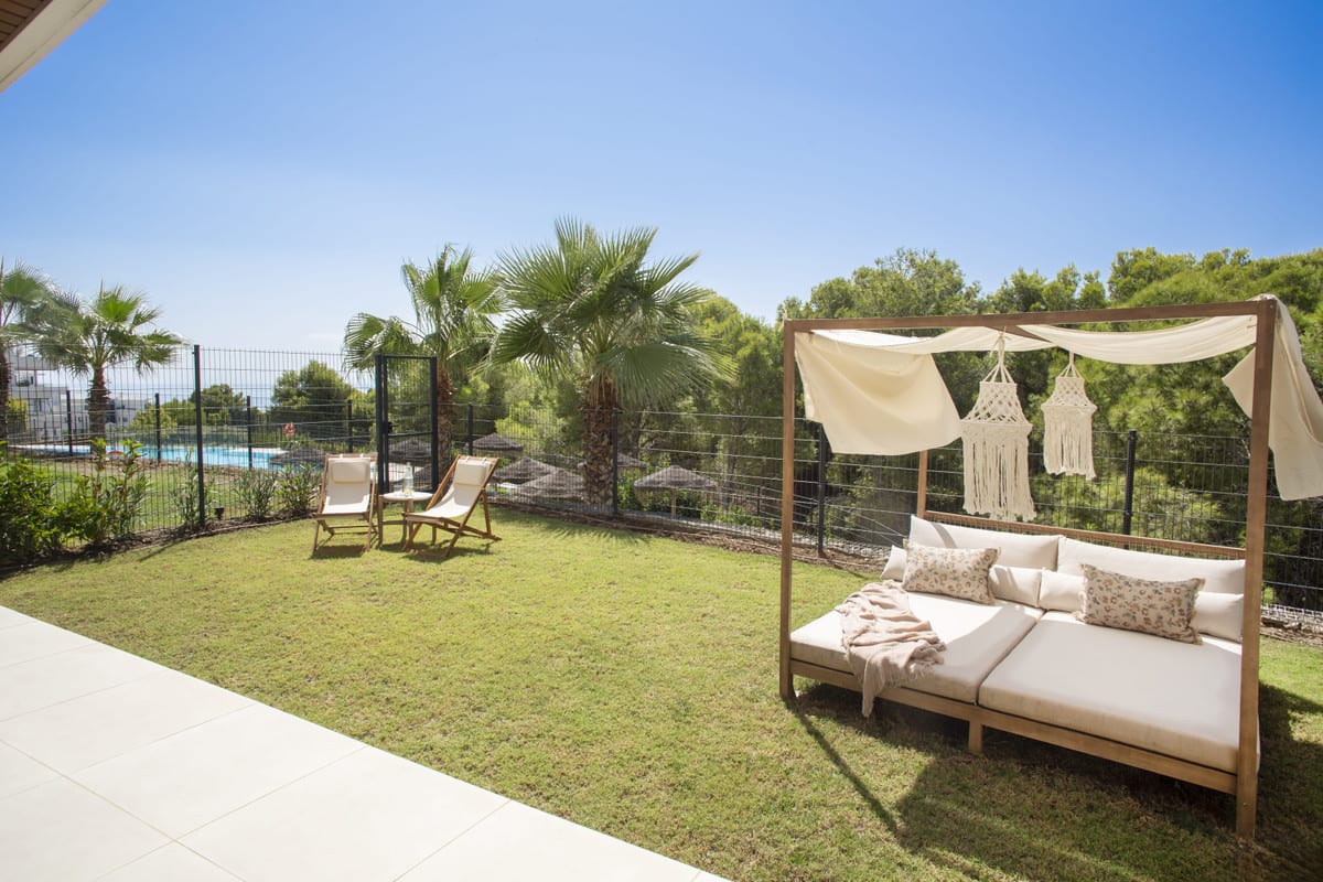 HIGUERON WEST with a lawn, wooden daybed with cream fabrics and a macramé hanging decoration, two folding chairs and palm trees.