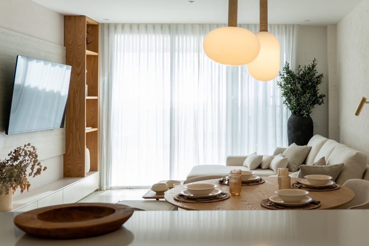 HORIZON36 interieur met gedekte houten eettafel, twee bolvormige hanglampen, crèmekleurige hoekbank, houten wandmeubel met televisie en witte vloergordijnen.