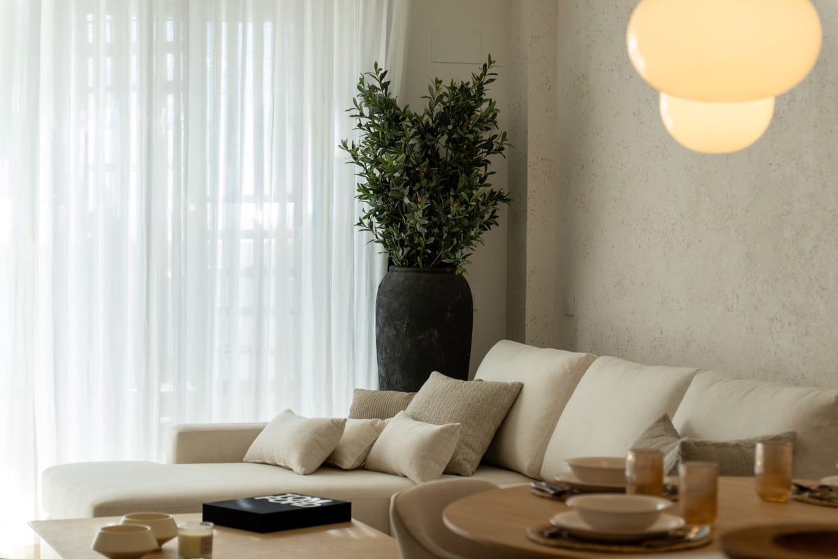 HORIZON36 detailopname van de zithoek met crèmekleurige bank, sierkussens, donkere plantenpot met groene plant, witte vloergordijnen en een gedeeltelijk zichtbare ronde eettafel op de voorgrond.