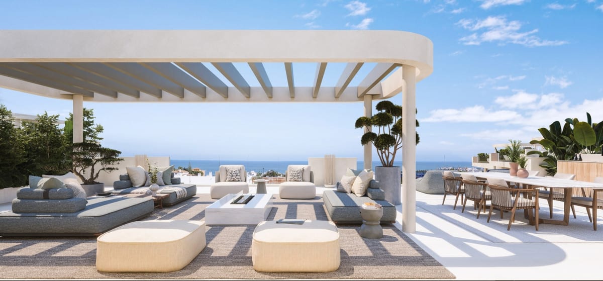 Rendered project visualisation of IDYLLIC with a spacious terrace beneath a concrete pergola, a lounge area with sofas and ottomans, a dining table on the right, large planters with trees and sea views in the background.
