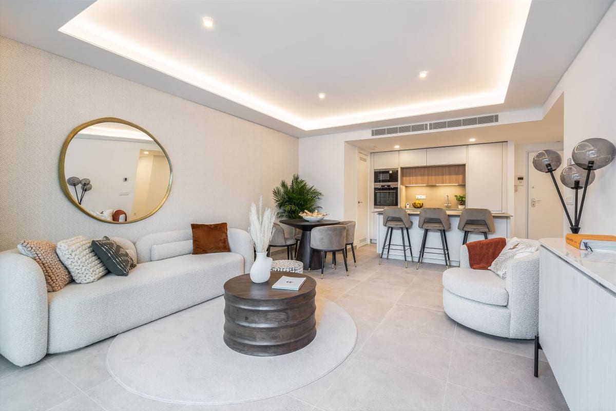 Living area at JADE TOWER with a light-coloured sofa, light-grey swivel armchair, dark cylindrical coffee table, round dining table with chairs and an open kitchen with bar stools in the background.