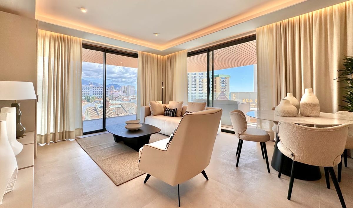Living and dining area at JADE TOWER with large sliding doors, beige curtains, a beige armchair in the foreground, light-coloured sofa, dining table with chairs and views of buildings and mountains outside.