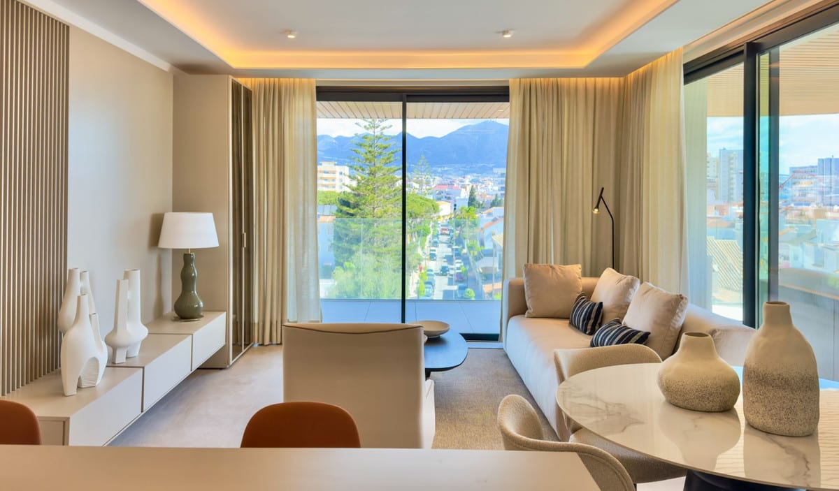 Living area at JADE TOWER with a beige sofa, low white cabinet, wooden wall slats and large sliding doors overlooking the street, trees and mountains outside.
