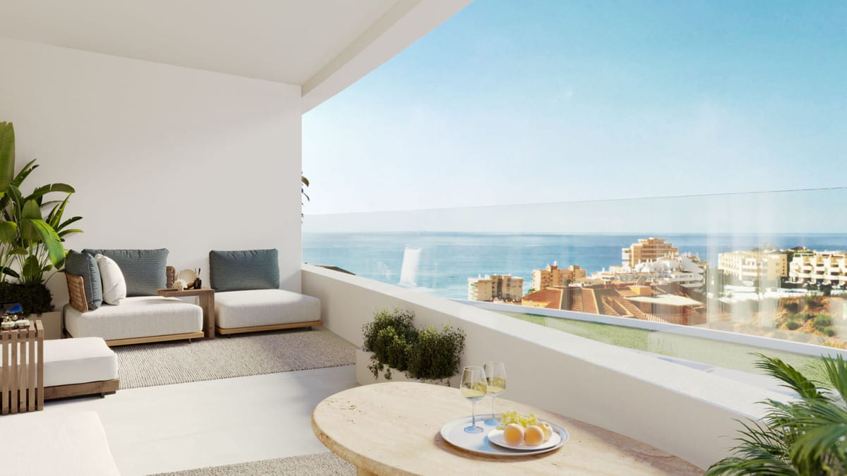 Project visualisation of KORALL with an open terrace, light lounge seating with blue cushions, a round marble table with glasses and fruit, tropical plants and a glass balustrade, with the sea and buildings visible in the distance.