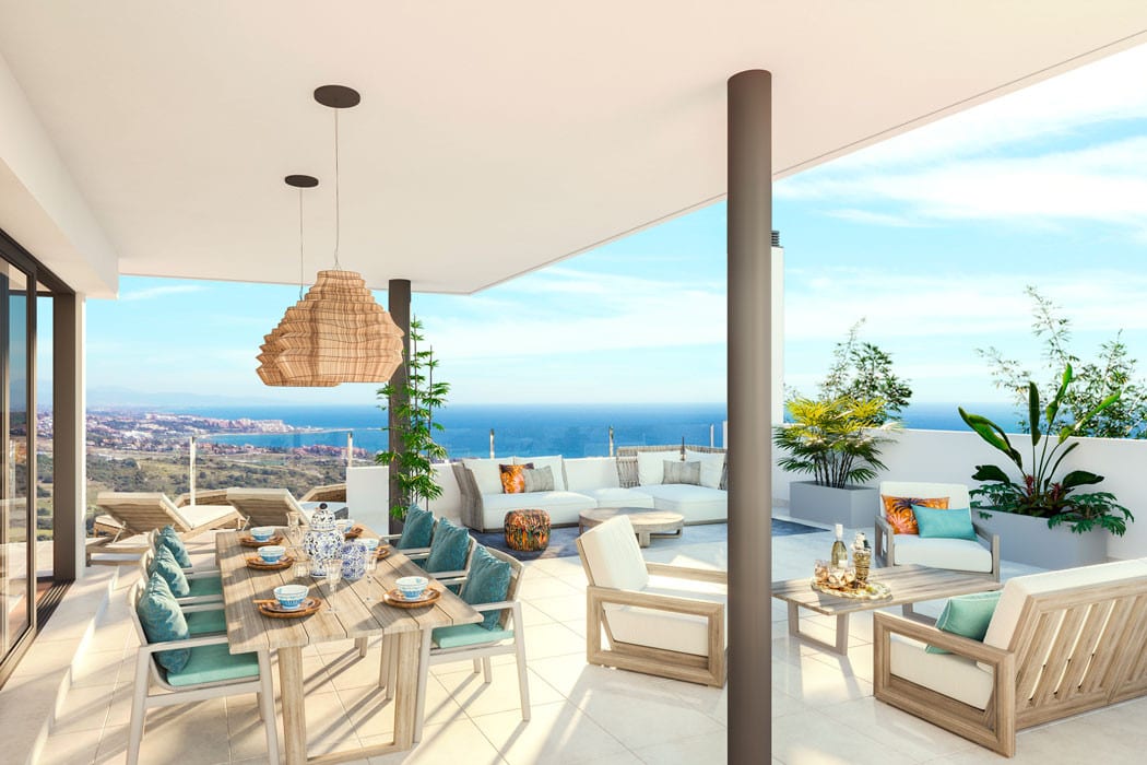 Project visualisation of the covered terrace at LAS TERRAZAS DE CORTESIN BON AIR with a wooden dining table, light seating area, low white balustrade and views of the sea and coastline.