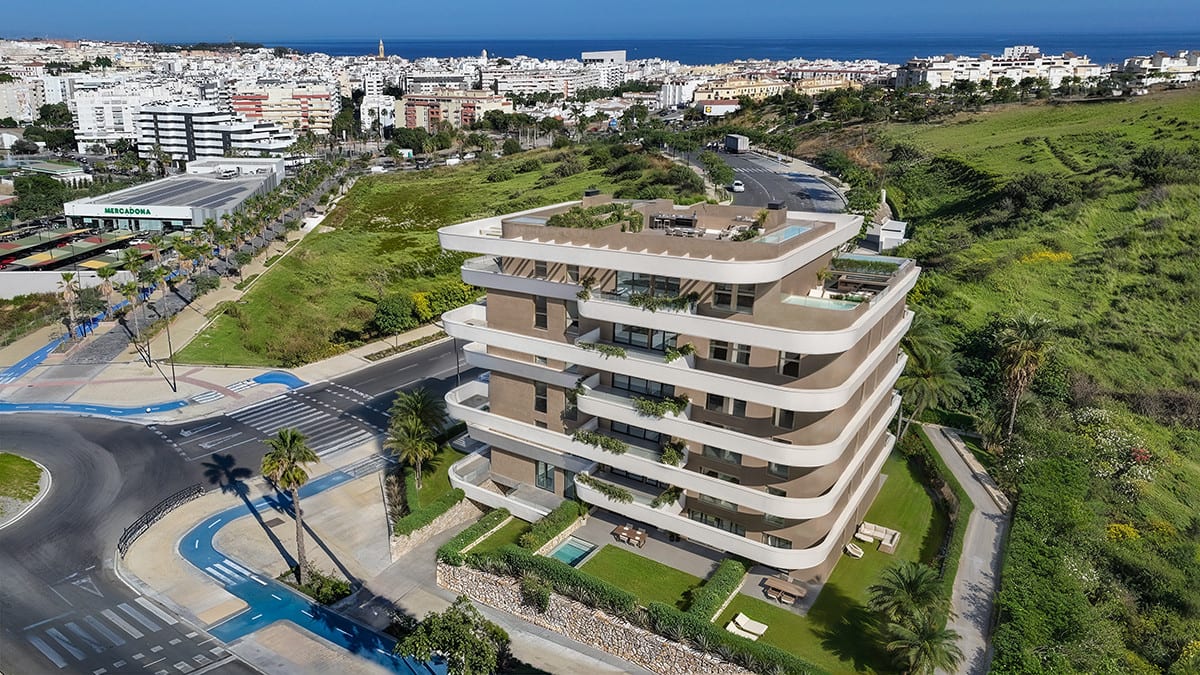 Aerial view of LOOA ESTEPONA showing the multi-storey building, a planted roof terrace, surrounding roads, green grounds and an urban panorama towards the coast in the background.
