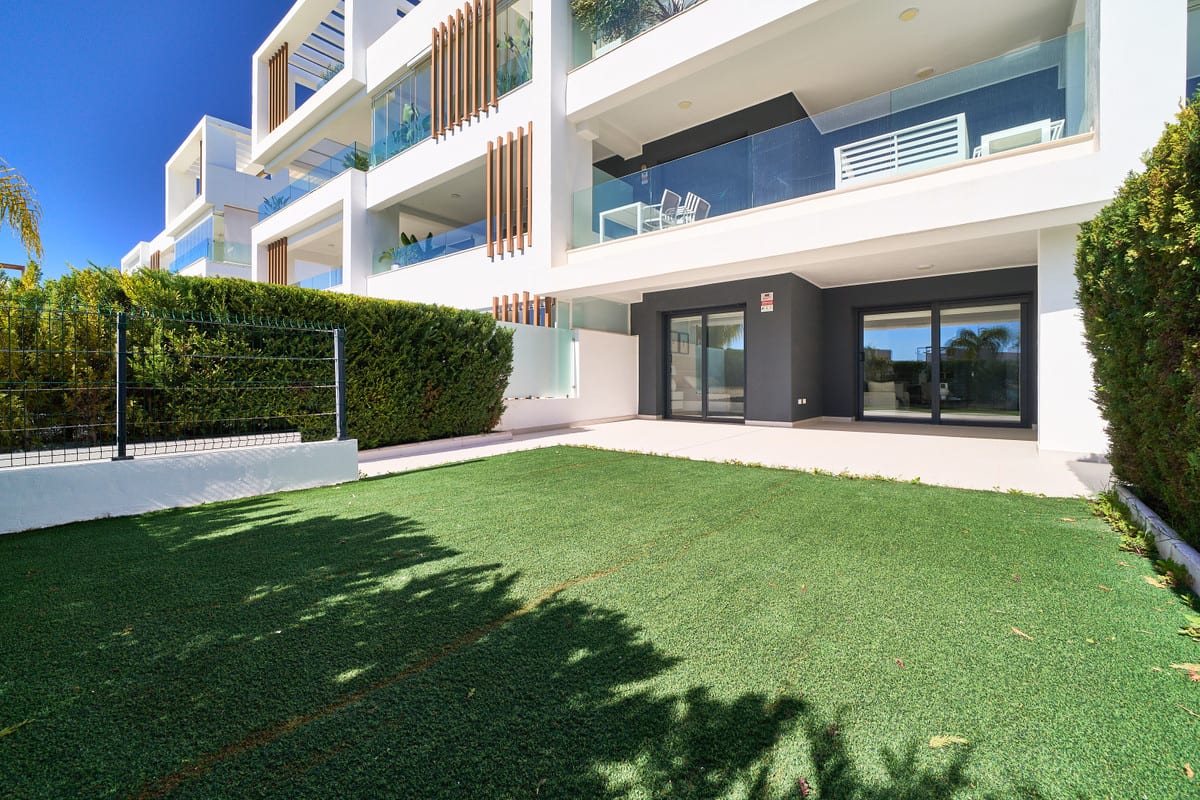 LOS MIRADORES DEL SOL with green lawn, light tiled terrace, dark sliding doors, glass balustrades and white façades with vertical wooden slats.