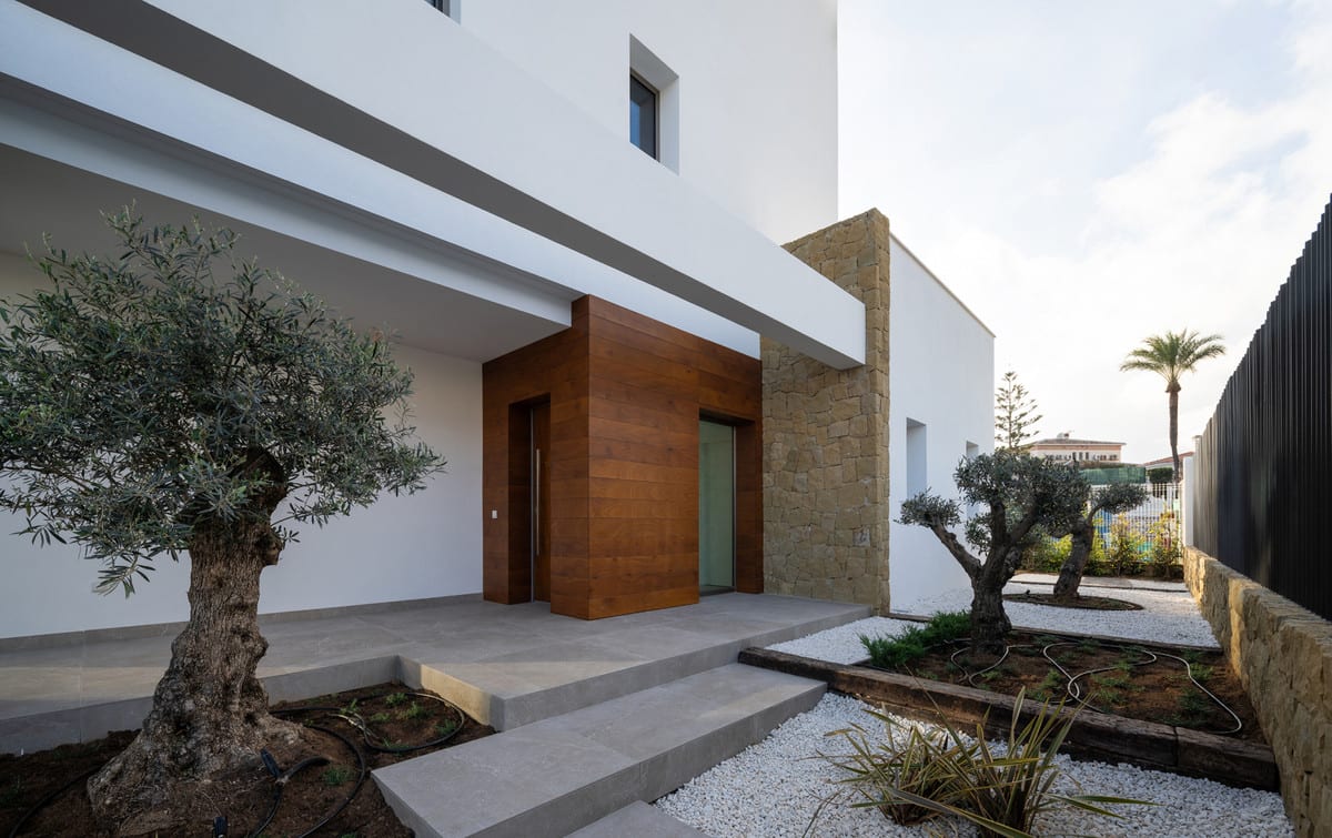The entrance combines a timber entrance volume with a natural-stone wall, wide grey steps and olive trees.