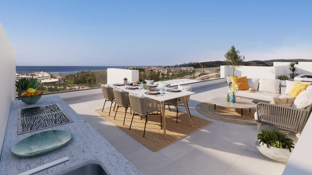 Project visualisation of MALA KAI featuring a spacious terrace, a laid dining table with woven chairs on a jute rug, a lounge area with yellow cushions, an outdoor kitchen worktop on the left, and views of the sea and wooded hills in the background.