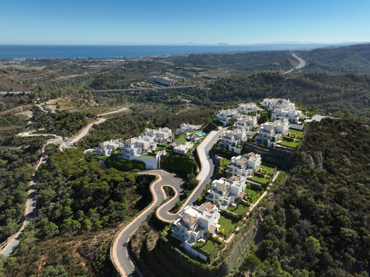 Aerial view of MARBELLA CLUB HILLS PHASE II with white buildings on a green hill, winding roads, a viaduct and the sea on the horizon.