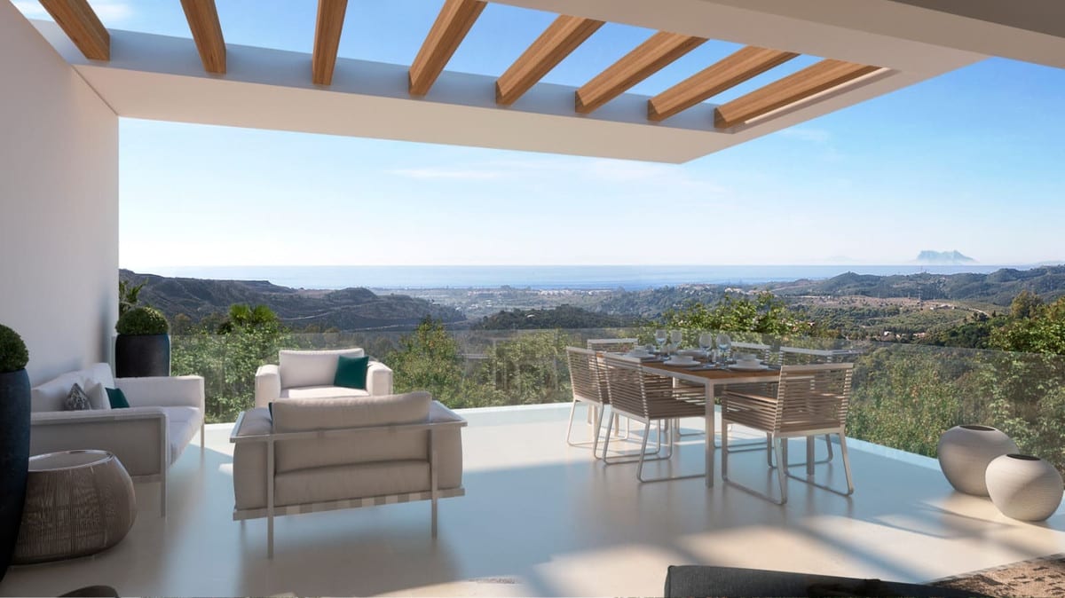 Project visualisation of MARBELLA CLUB HILLS PHASE II with a covered terrace, wooden beams, light-coloured seating, a dining table and a glass balustrade overlooking hills and the sea.