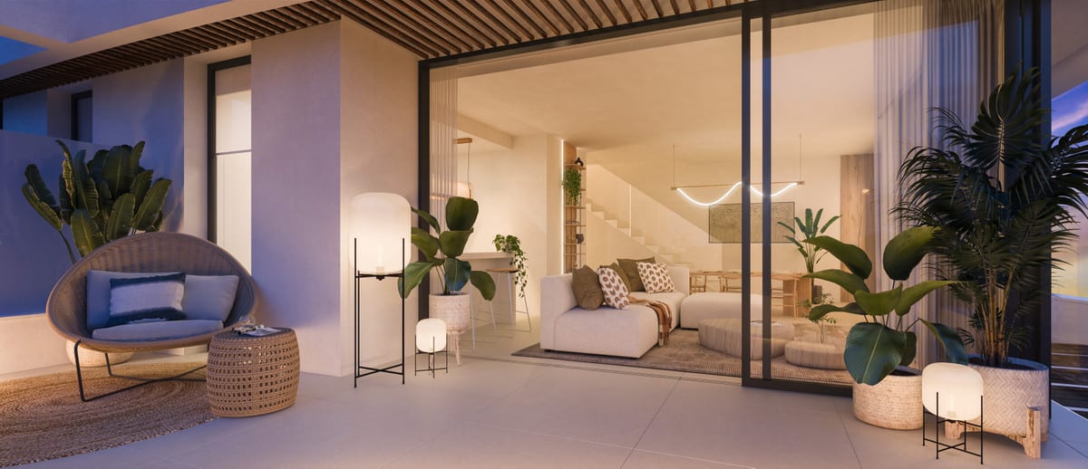 Evening project visualisation of MARINA GOLDEN BAY featuring an illuminated terrace, a rattan armchair, woven side table, large tropical plants and a warmly lit living room visible behind sliding glass doors.
