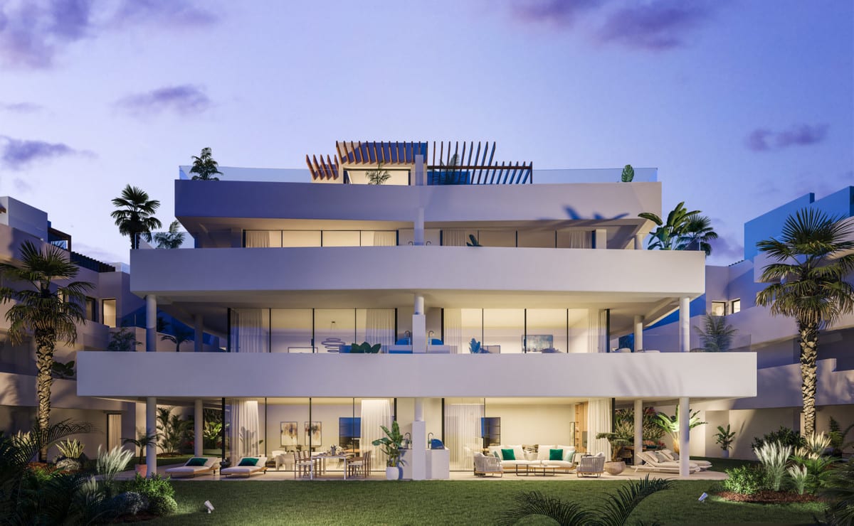 Project visualisation of MIRADOR DE ESTEPONA HILLS featuring a three-storey white building, wide balconies, glass sliding doors, a wooden pergola on top and landscaped planting at dusk.